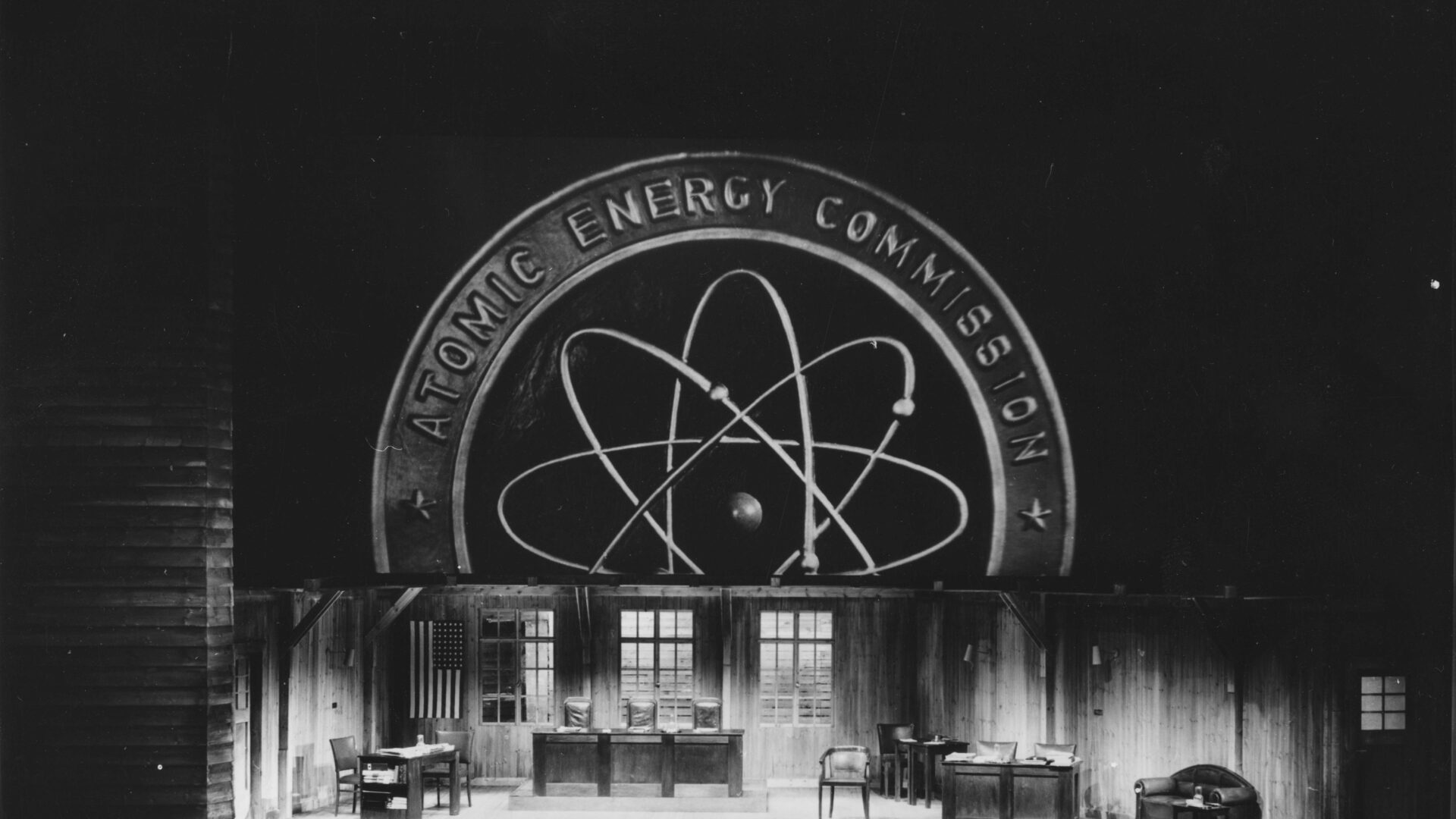 Rießiges Atomic Energy Commission Logo im Hintergrund, Tischen und Stühlen auf der Bühne davor