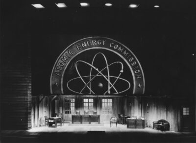 Rießiges Atomic Energy Commission Logo im Hintergrund, Tischen und Stühlen auf der Bühne davor