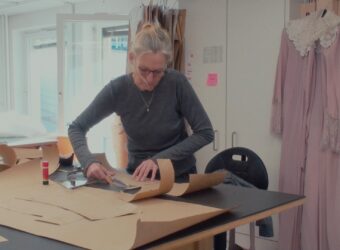 Gabriele Behne steht an einem Tisch im Atelier, schneidet Schnittmusterteile für Kostüme, im Hintergrund hängen Kostümteile.