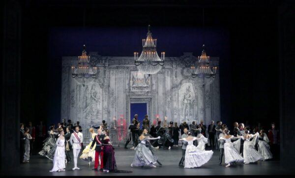 Tanzende Ball-Gäste auf einer Bühne vor eindimensionalem Bühnenhintergrund, darüber drei Kronleuchter